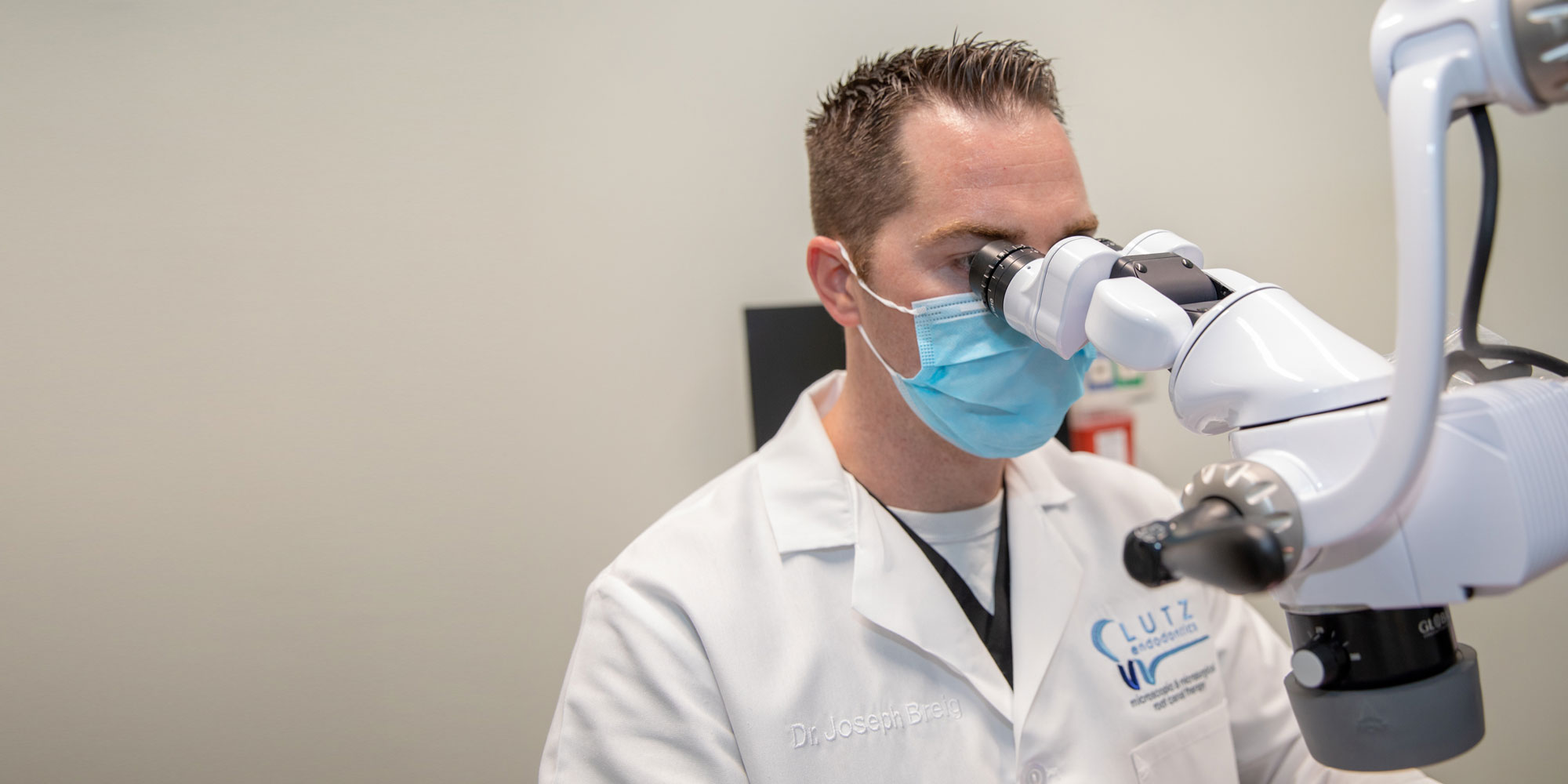 A man wearing a lab coat and mask looking through a microscope.